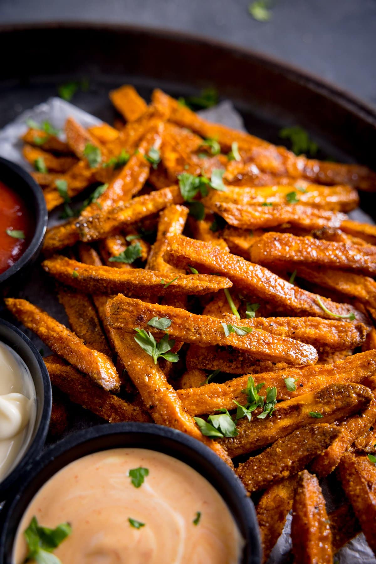 A close up of cooked sweet potato fries, sprinkled with parsley, with some dips in small bowls on the side