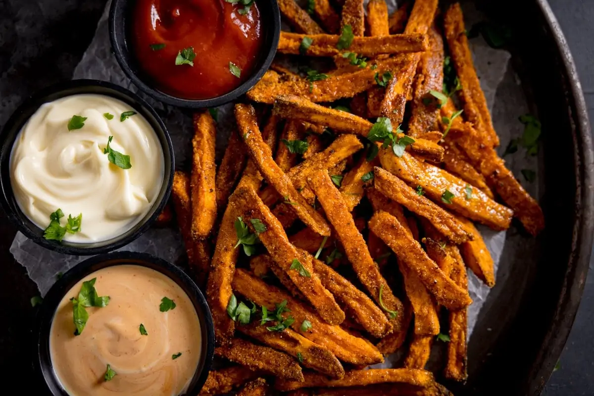 Cooked sweet potato fries, sprinkled with chopped parsley, with three dips in small bowls alongside on a greaseproof paper background.