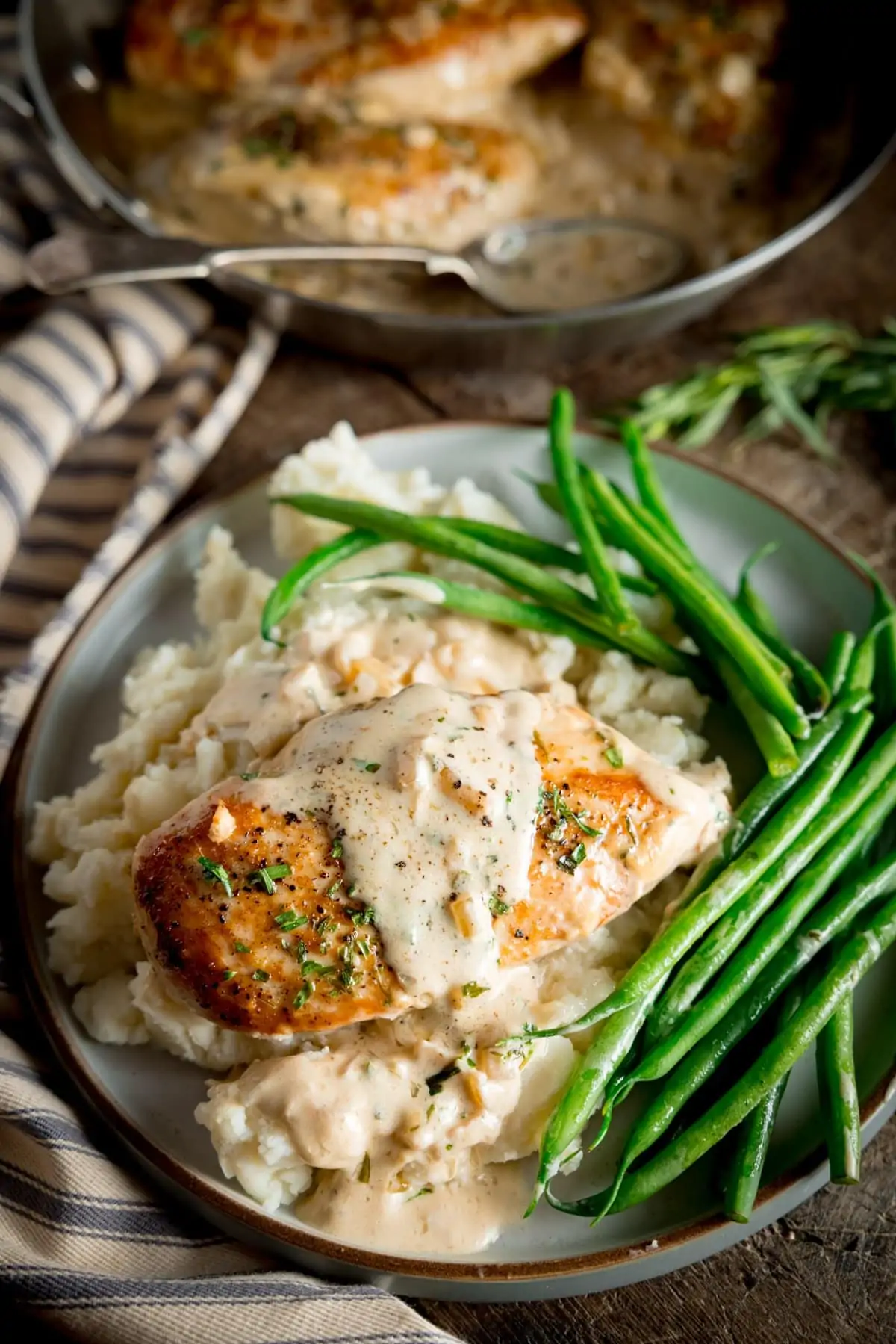 A finished plate of Tarragon chicken, mashed potato and green beans with sauce over the top, set on a stripy tea towel with a bowl of mashed potato to the side