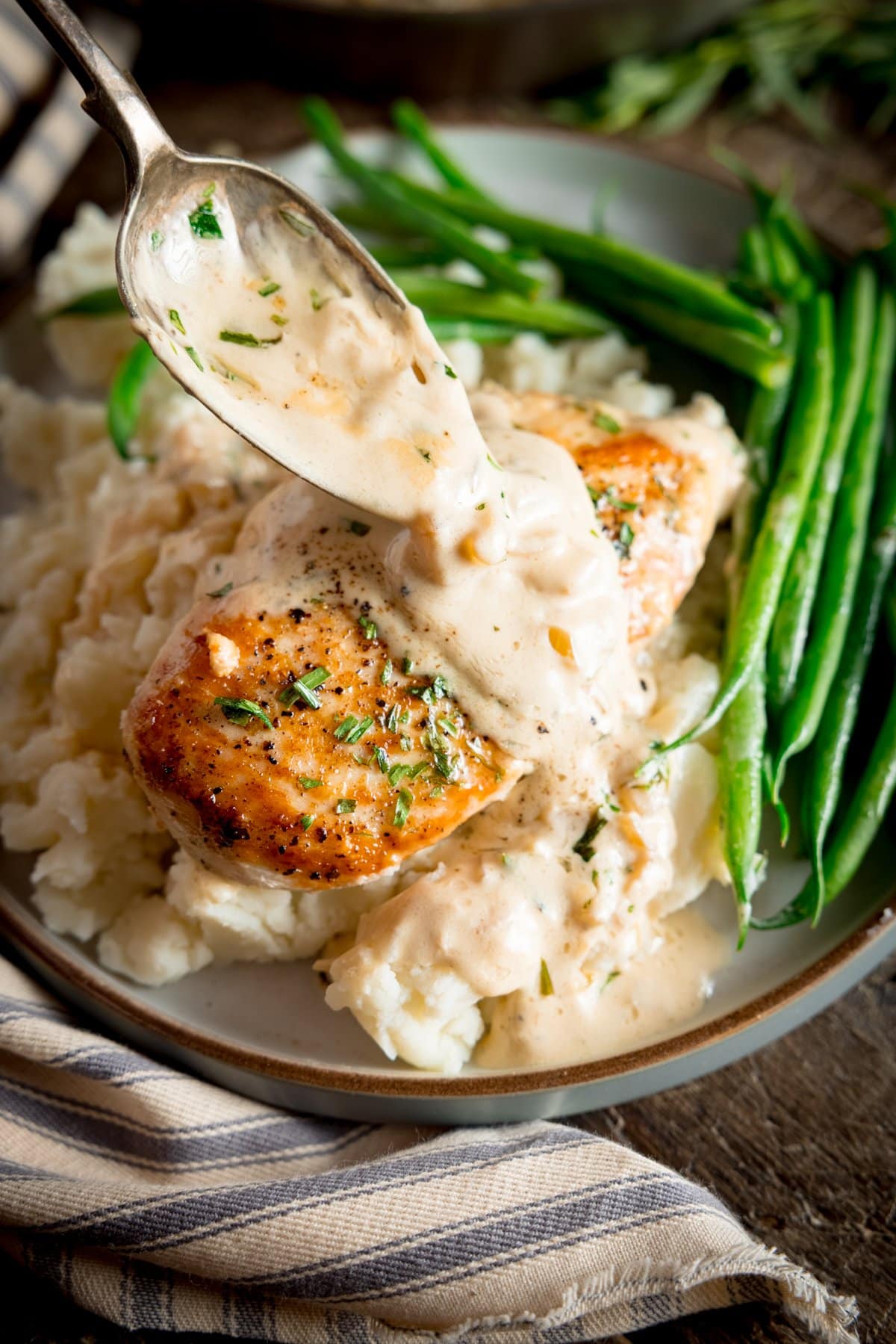 A light grey plate with mashed potato, green beans and creamy Tarragon Chicken on top, with a spoon drizzling the creamy sauce over the top