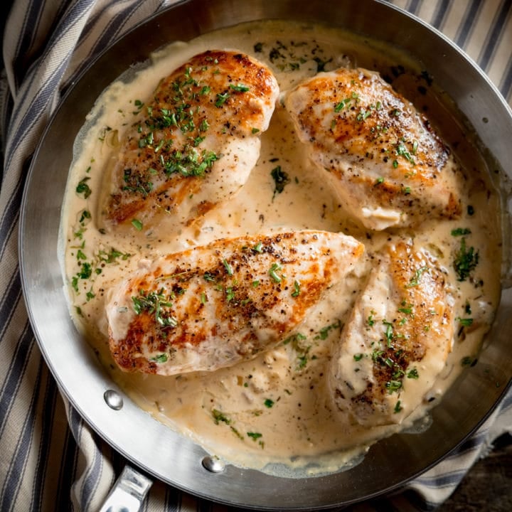 A square photo of a pan of cooked Tarragon chicken in a creamy sauce, sprinkled with chopped parsley, laid on top of a stripy grey and white tea towel