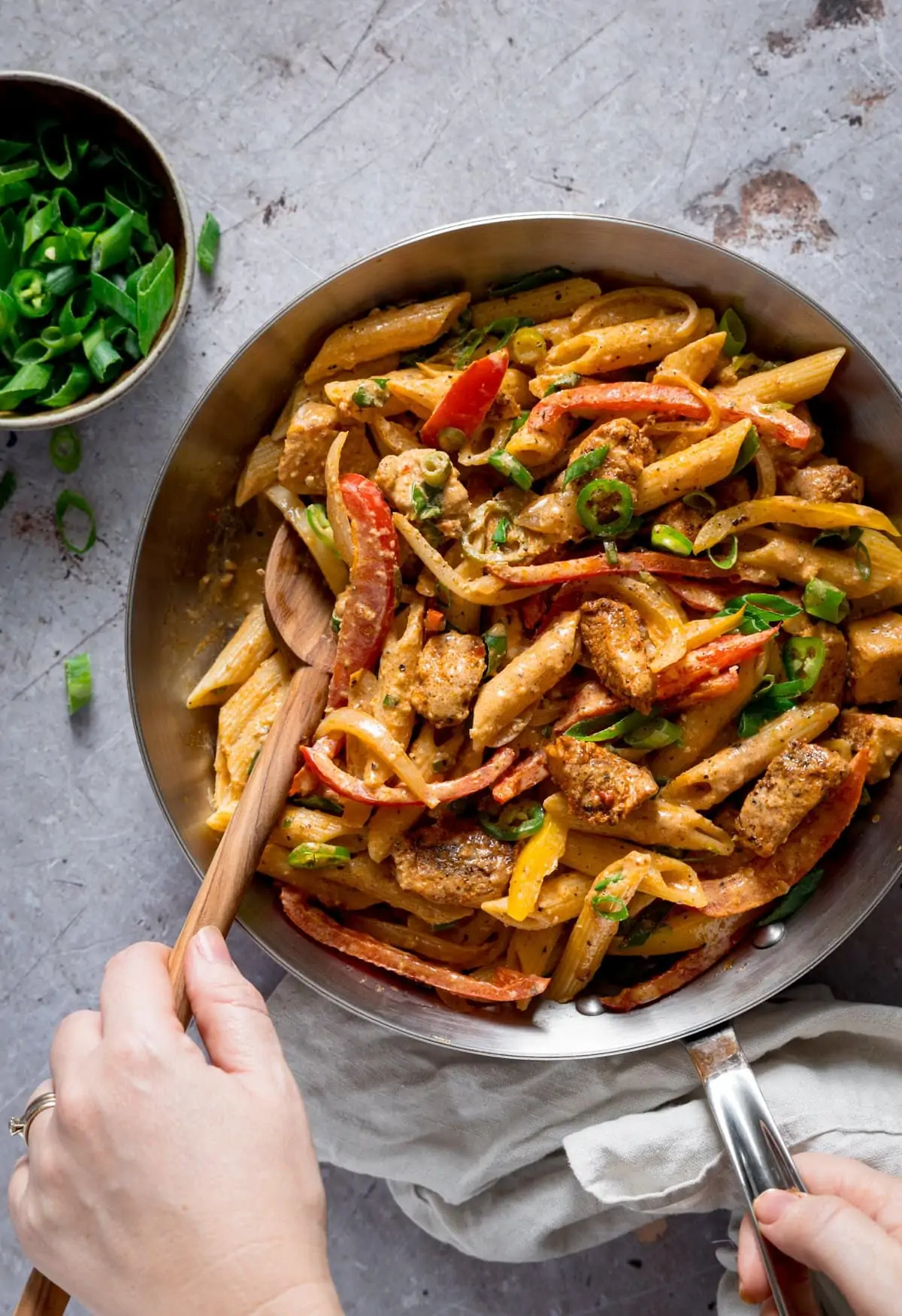 Buffalo pasta in a silver pan, being stirred, using a wooden spoon. The pan is on a light grey surface.