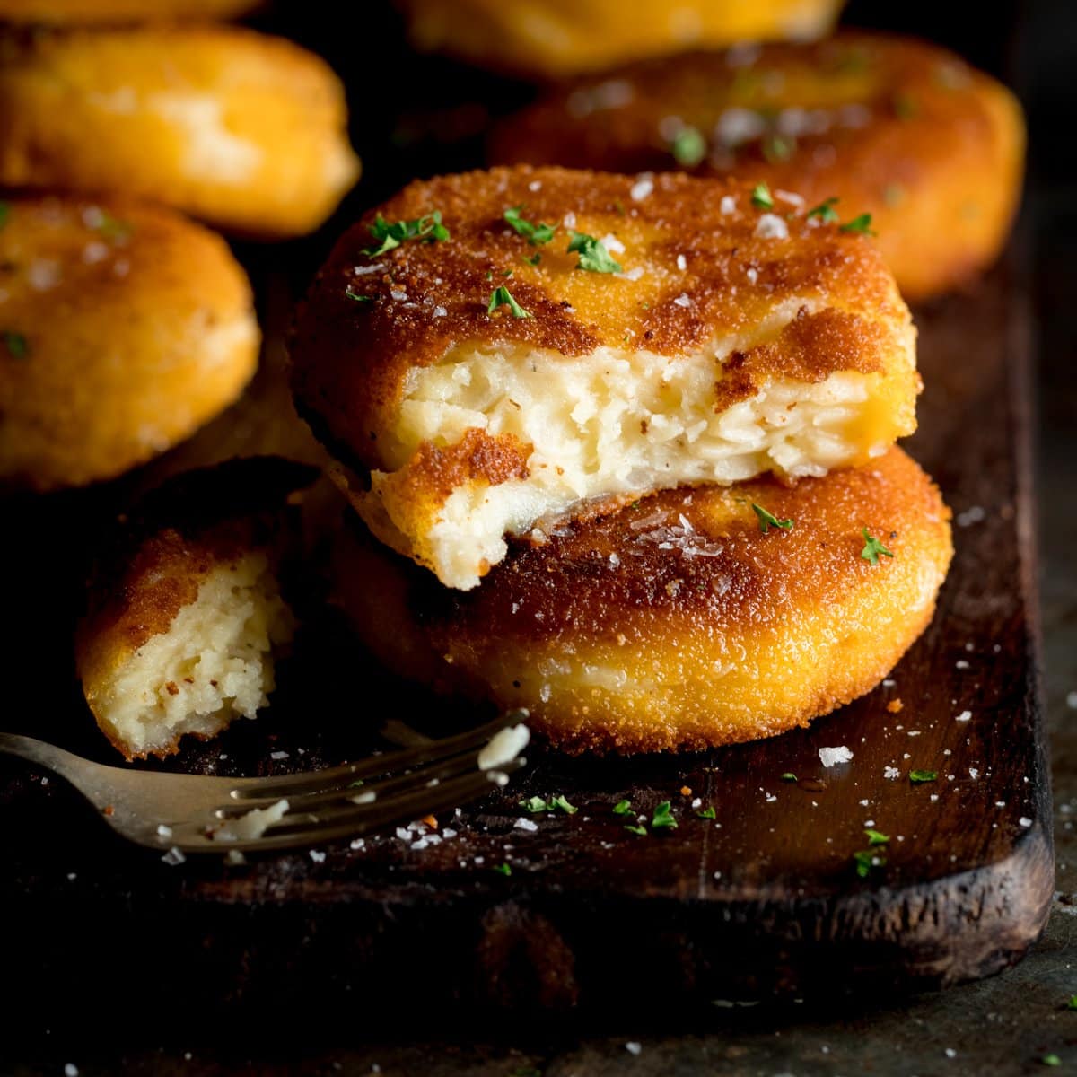 A wooden board with cheesy potato cakes piled up and one sliced in half sprinkled with chopped parsley