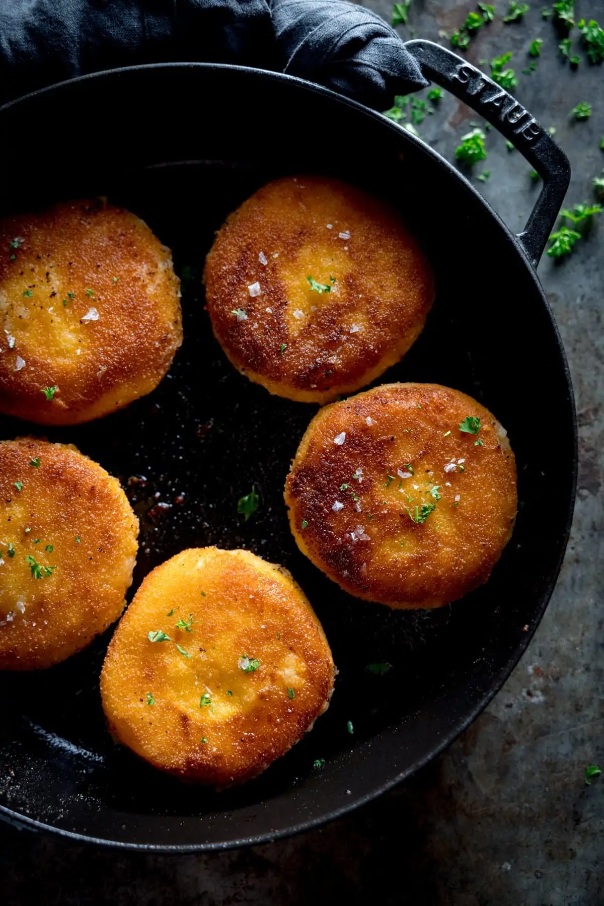 A black pan full of cheesy potato cakes all golden brown and crispy, sprinkled with parsley on a grey table