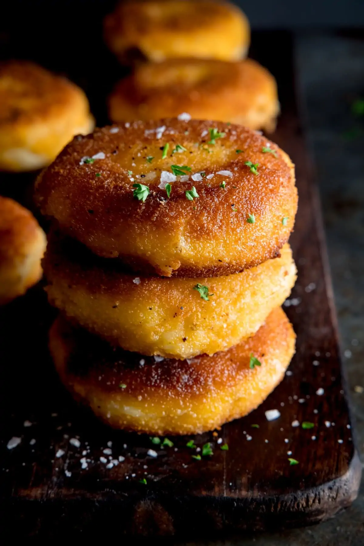 A little pile of three cooked cheesy potato cakes with more in the back ground, all sprinkled with salt and chopped parsley