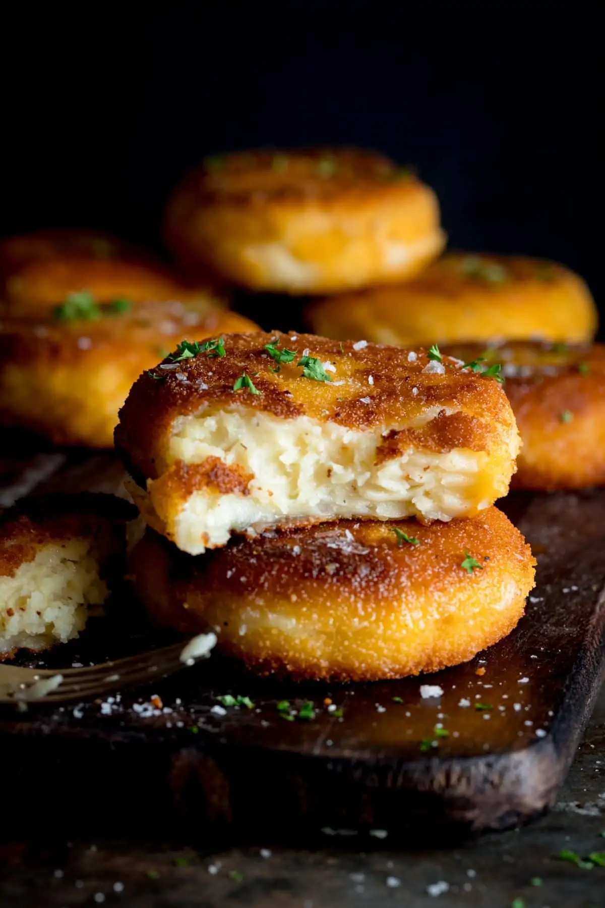 A wooden board full of cooked cheesy potato cakes, sprinkled with salt and chopped parsley, one is cut in half and oozy.