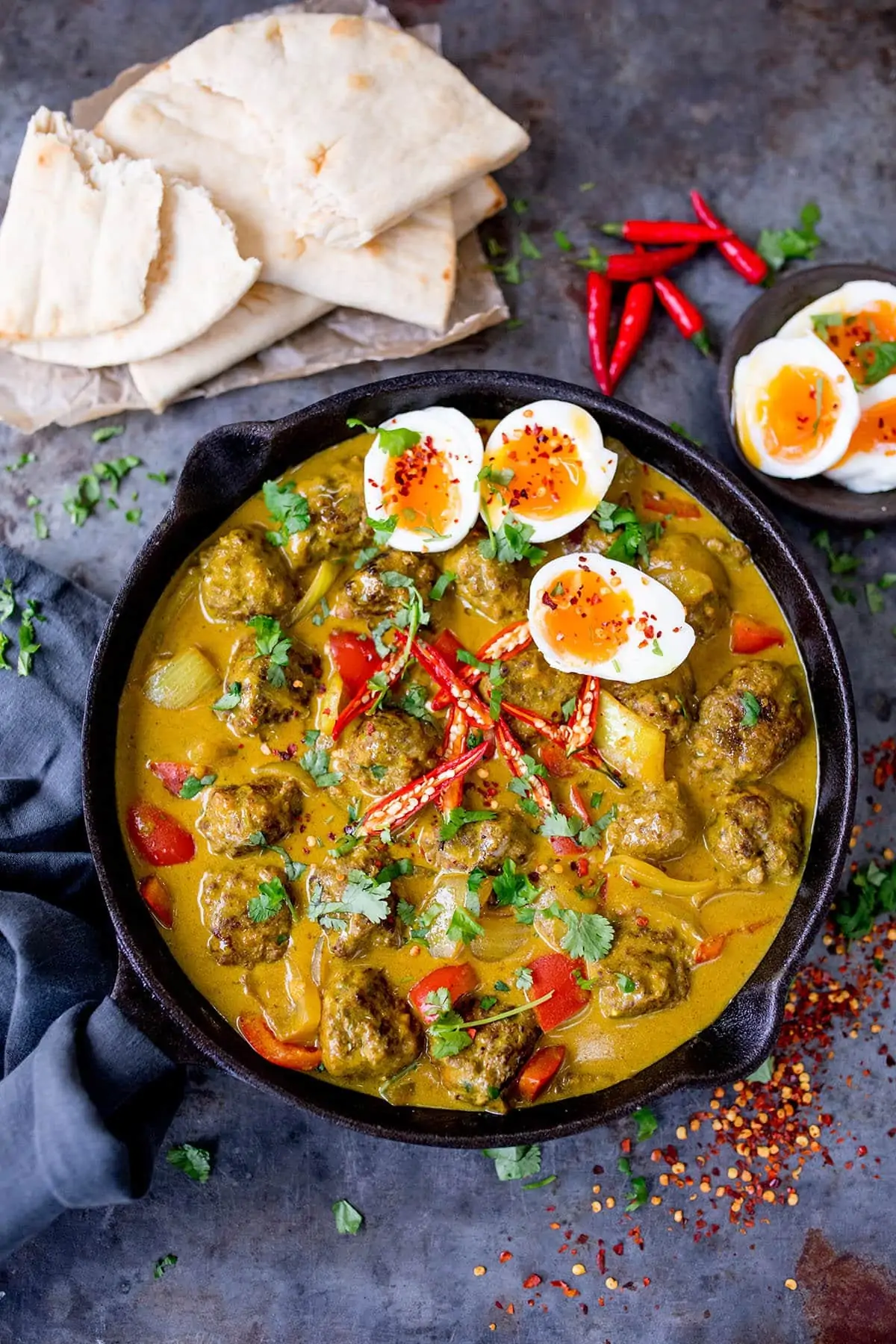 Chinese style meatball curry in a black cast iron pan on a blue board with some pitta breads and boiled eggs in the background.