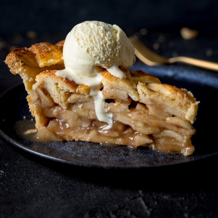 A grey plate with a slice of deep dish apple pie on top with a scoop of vanilla ice cream on the top with a napkin and fork in the background on a grey table.