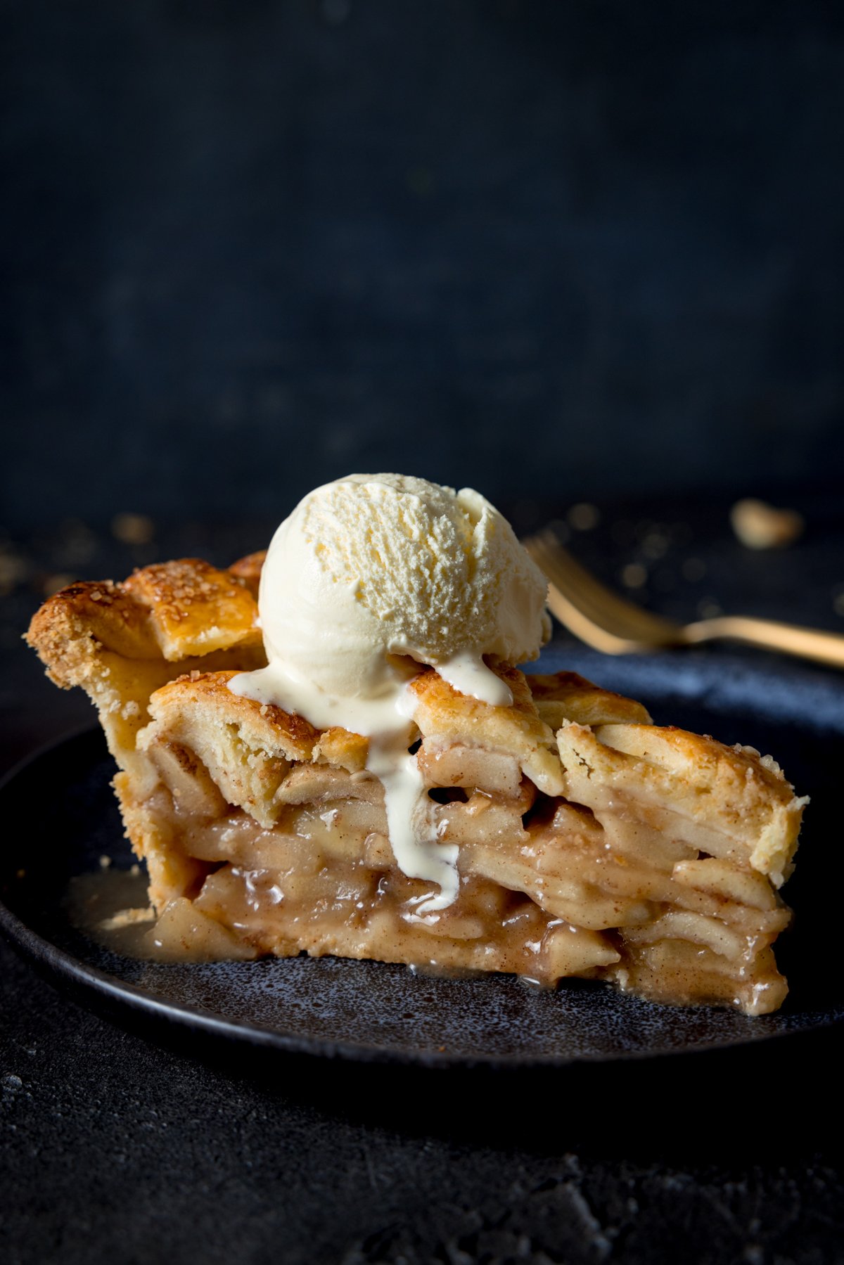 A side shot of a slice of deep dish apple pie with a scoop of vanilla ice cream on top, on a blue plate with a napkin and fork in the background on a grey table.