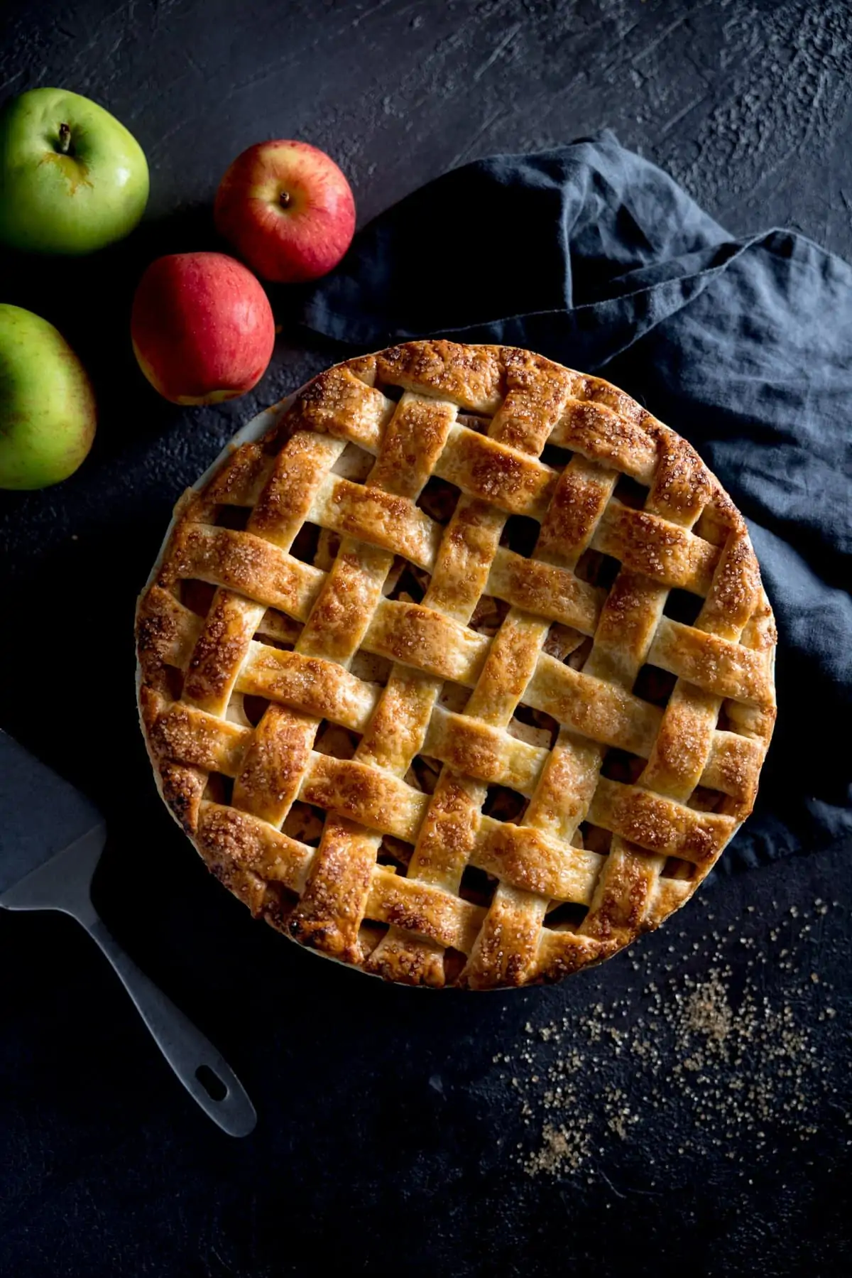 A photo looking down on a perfectly round golden brown lattice topped deep dish apple pie, with apples in the back ground, a blue tea towel and and a spoon next to the dish.