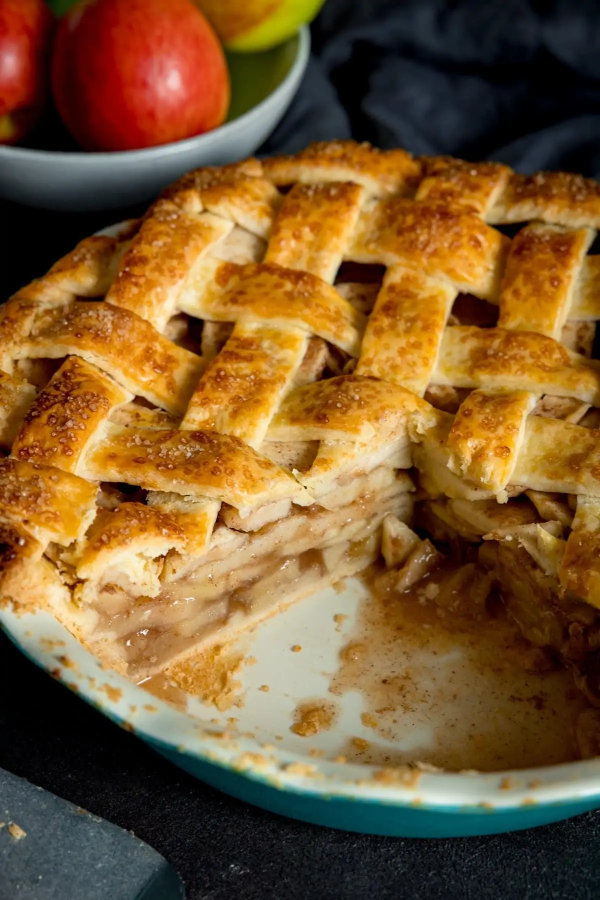 A big round lattice topped deep dish apple pie cooked and golden brown with a wedge cut out of it, in a white tin