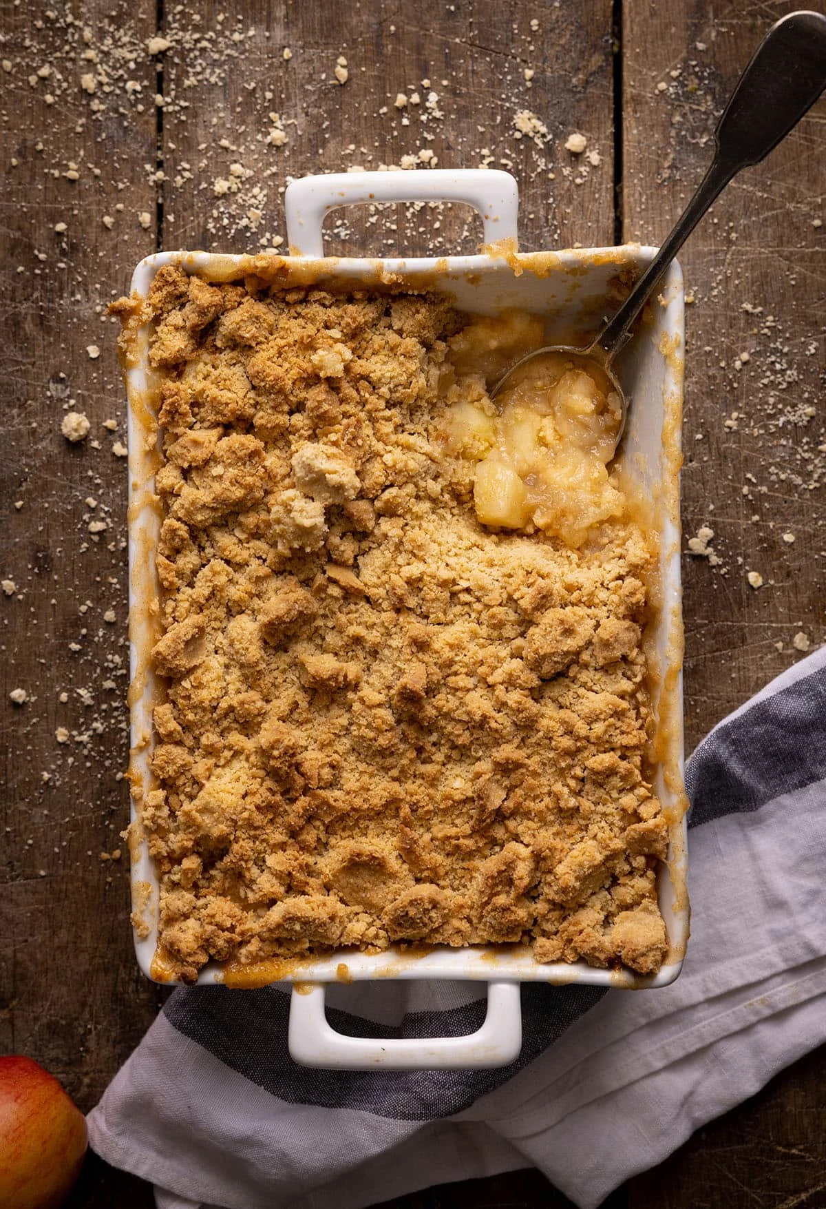 Overhead picture of a baking dish full of apple crumble with a spoon in it sat on a rustic wooden table.