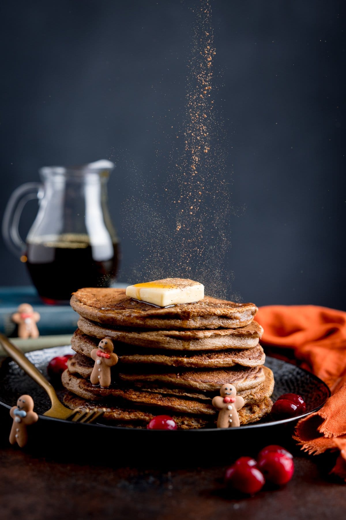 Edible glitter being sprinkled onto a stack of gingerbread pancakes with a jug of maple syrup in the background.
