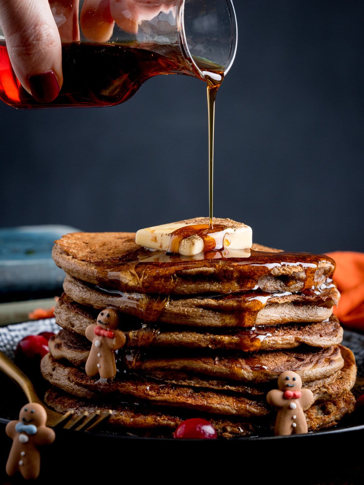 Maple syrup being poured over a large stack of gingerbread pancakes with mini gingerbread men on the plate along with some fresh cranberries.