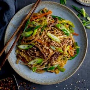 Square image of beef chow mein on a blue plate, with chopsticks resting on the plate.