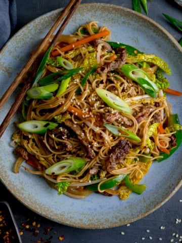 Square image of beef chow mein on a blue plate, with chopsticks resting on the plate.