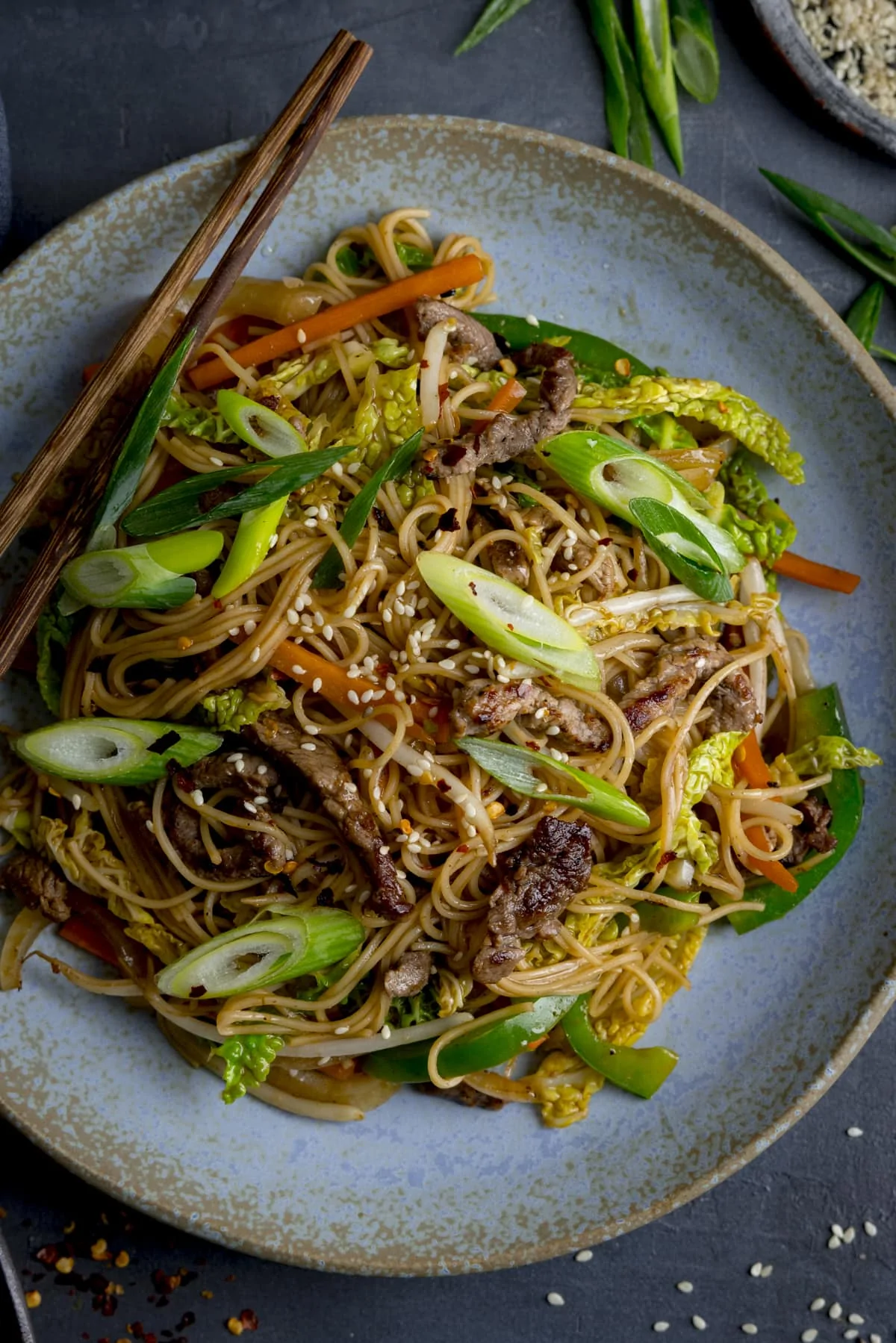 Beef chow mein on a blue plate on a grey background. There are chopsticks on the edge of the plate.