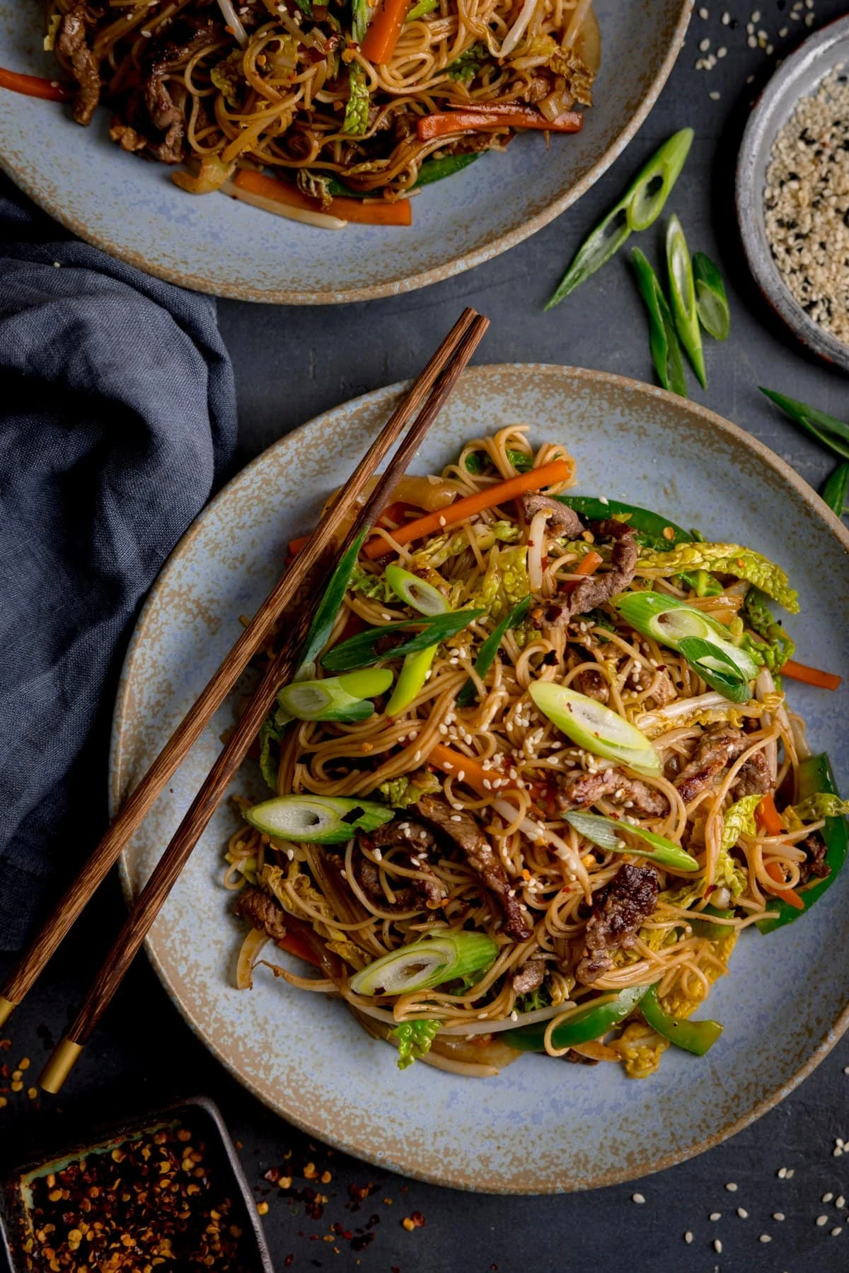 Tall image of beef chow mein on a blue plate with a set of chopsticks on top. The is a further plate of chow mein, just out of shot. The plates are on on a grey background with a napkin and some sesame seeds and spring onions around the dish.