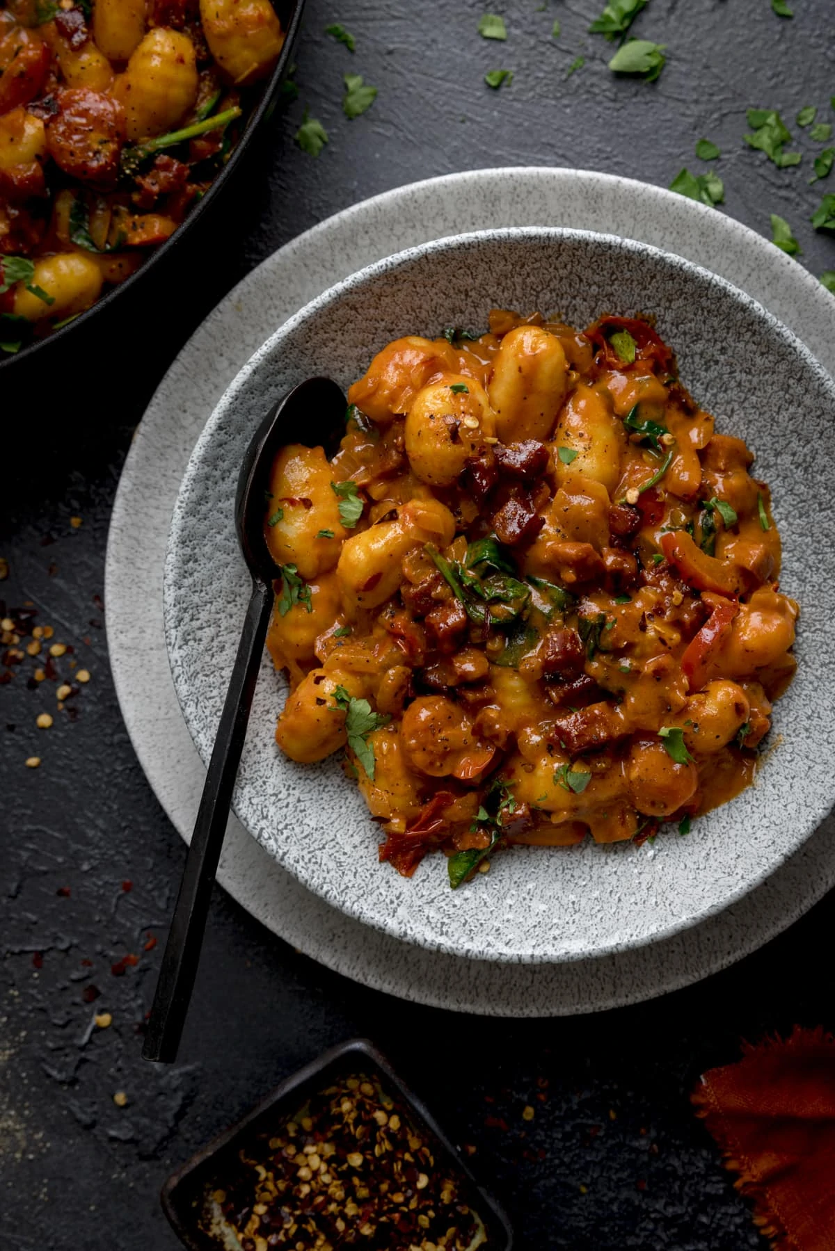 Gnocchi and chorizo in a grey bowl with a black spoon in. The bowl is on a grey surface and the pan is just in shot.