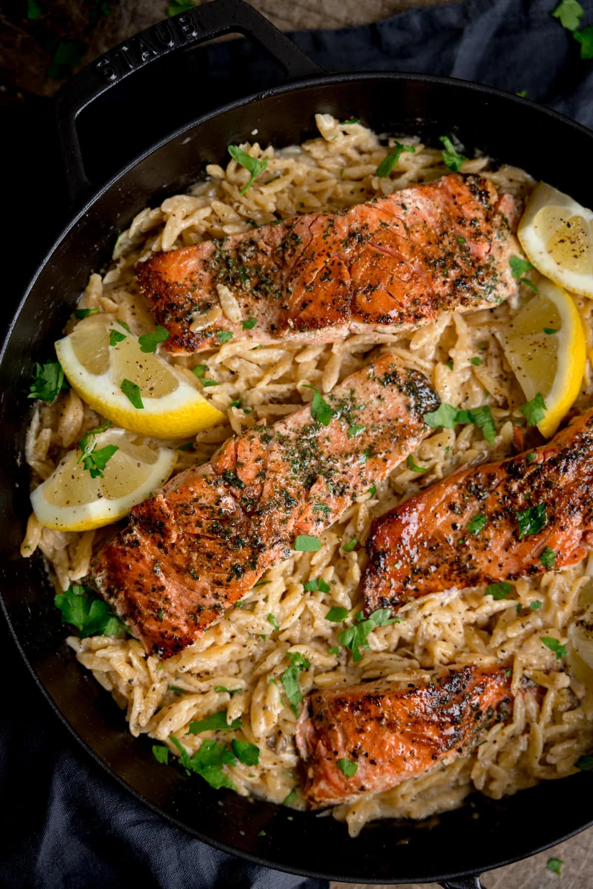 Overhead image of pan-fried salmon fillets nestled in a bed of creamy lemon orzo topped with parsley. There are lemon wedges at the edges of the pan.