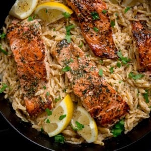 Square image of pan-fried salmon fillets nestled in a bed of creamy lemon orzo topped with parsley. There are lemon wedges at the edges of the pan.