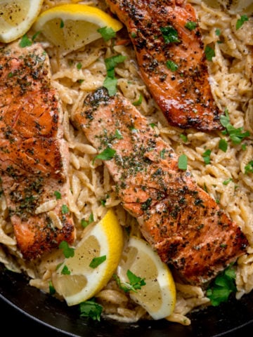 Square image of pan-fried salmon fillets nestled in a bed of creamy lemon orzo topped with parsley. There are lemon wedges at the edges of the pan.