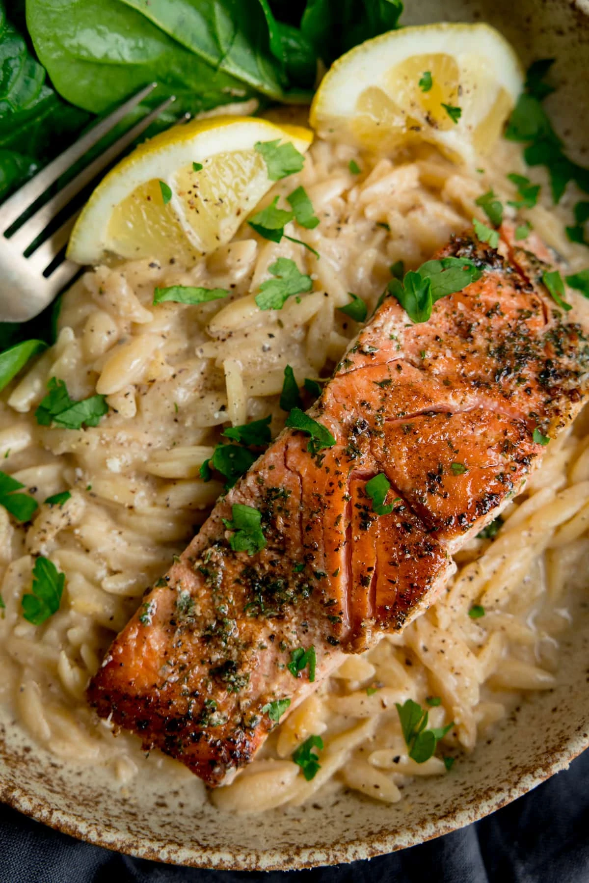 Pan-fried salmon fillet in a bowl with creamy orzo, lemon wedges and fresh spinach. There is a fork in the bowl too.