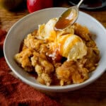 A white bowl filled with apple crumble topped with two scoops of vanilla ice cream. Caramel sauce is being drizzled over with a spoon. The bowl is on a wooden table next to a dark orange napkin. There are a couple of apples next to the bowl.