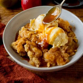 A white bowl filled with apple crumble topped with two scoops of vanilla ice cream. Caramel sauce is being drizzled over with a spoon. The bowl is on a wooden table next to a dark orange napkin. There are a couple of apples next to the bowl.