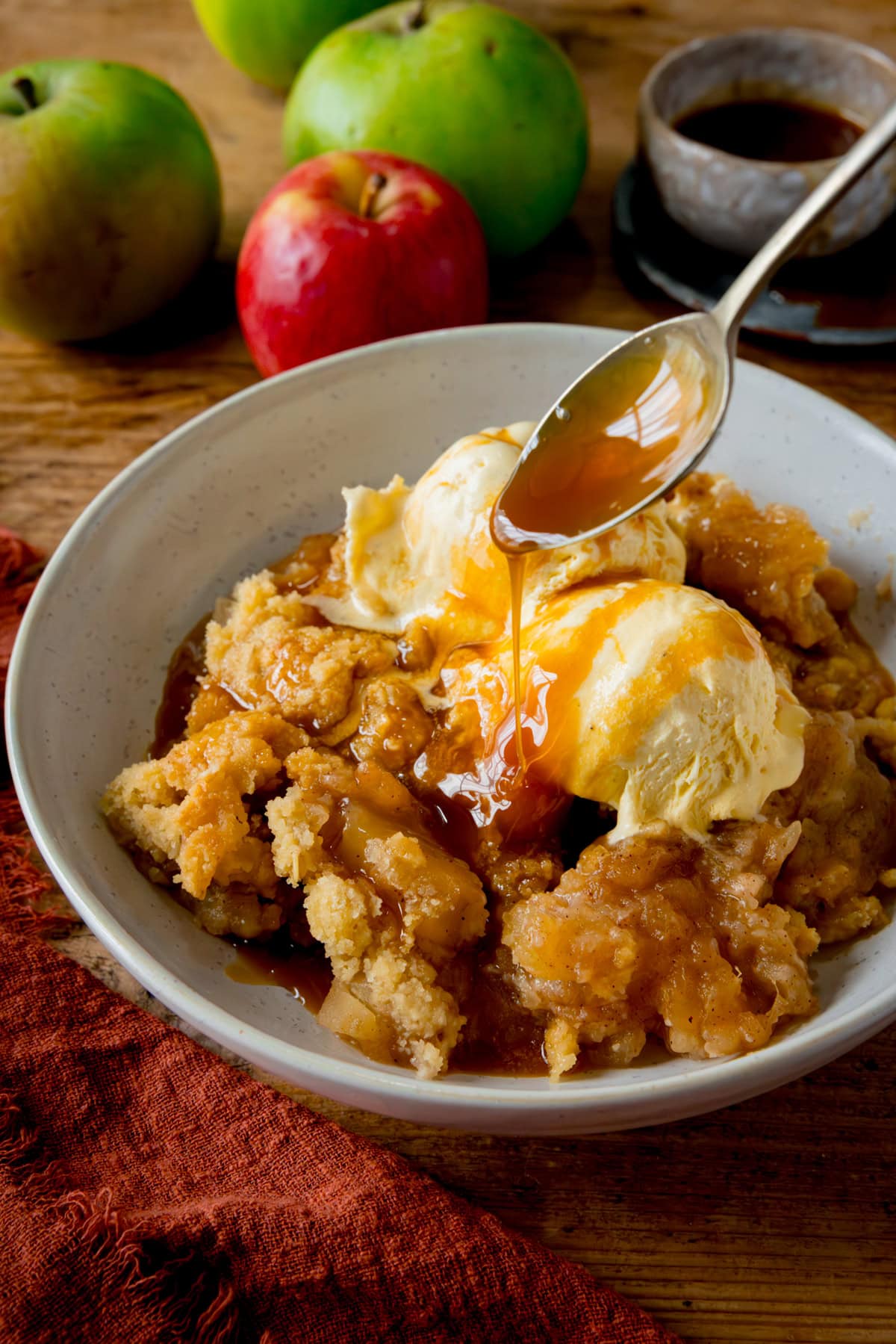 Tall image of a white bowl filled with apple crumble topped with two scoops of vanilla ice cream. Caramel sauce is being drizzled over with a spoon. The bowl is on a wooden table next to a dark orange napkin. There are a few apples and a small bowl of caramel sauce nearby.