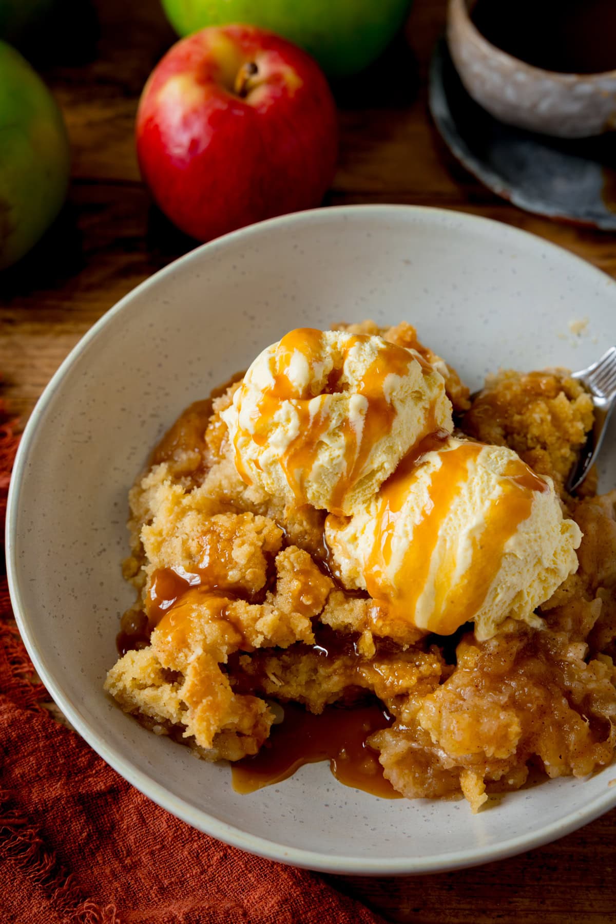 Tall image of a white bowl filled with apple crumble topped with two scoops of vanilla ice cream. Caramel sauce is drizzled over the ice cream. The bowl is on a wooden table next to a dark orange napkin. There are a few apples and a small bowl of caramel sauce nearby.
