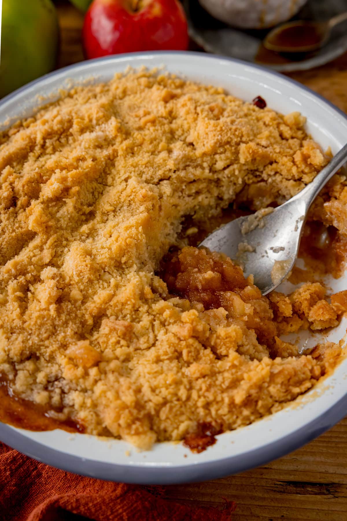 A baked apple crumble in a white dish with a scoopful taken out sits on a wooden table. There is a silver serving spoon scooping some of the apple filling from the dish.
