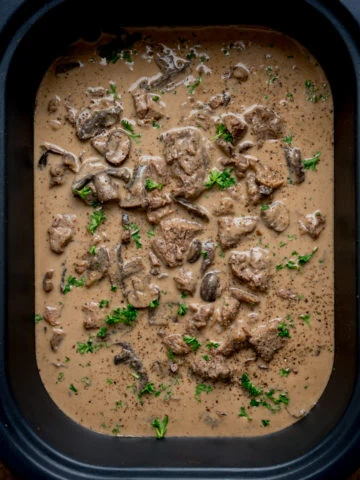 Overhead square image of slow cooked beef stroganoff, with parsley on top, in a slow cooker, on a wooden table.
