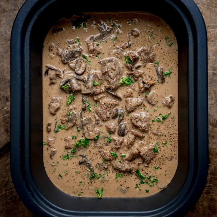 Overhead square image of slow cooked beef stroganoff, with parsley on top, in a slow cooker, on a wooden table.