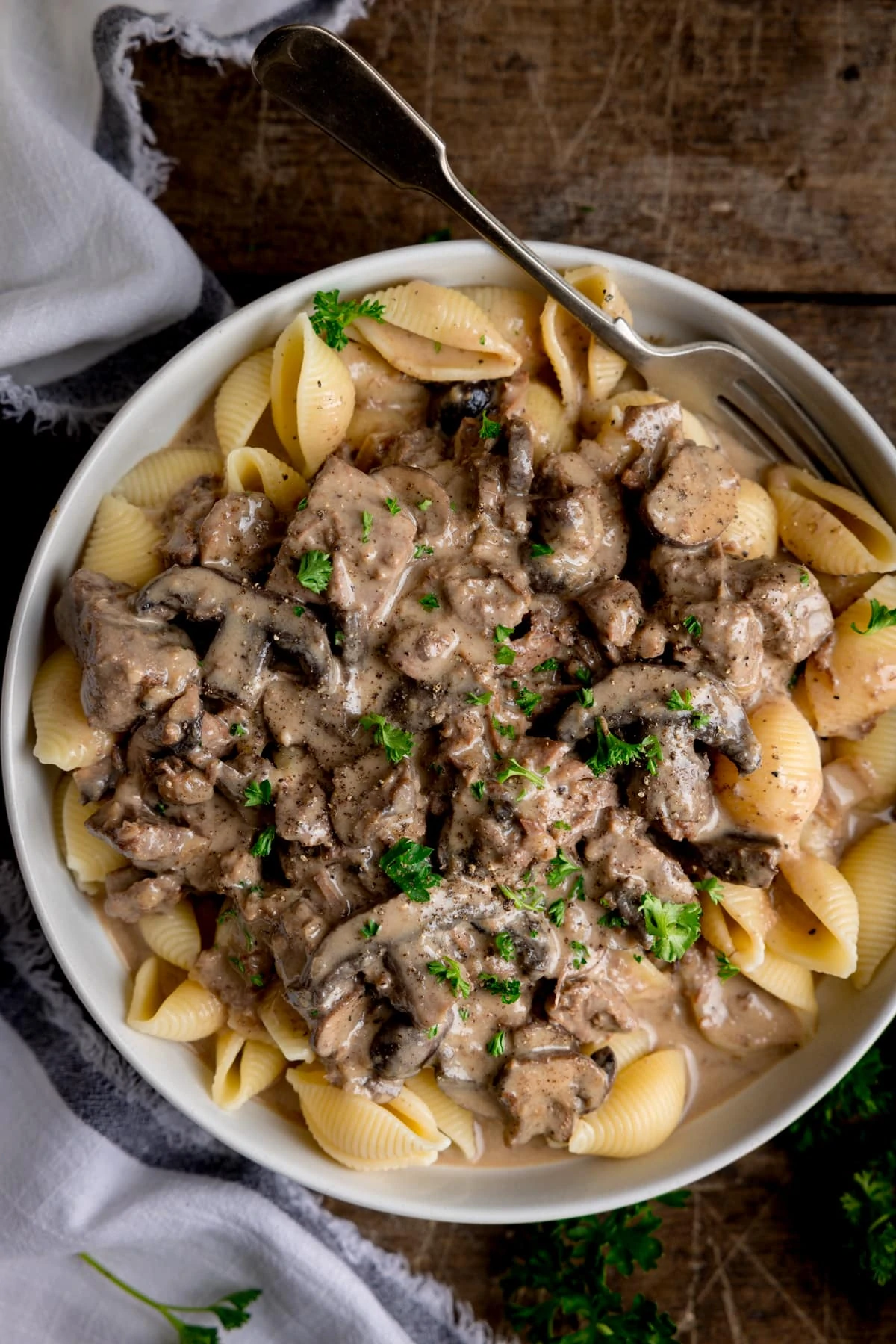 White bowl filled with slow cooker beef stroganoff served over pasta shells. Parsley is sprinkled on top. The bowl has a fork in it. The bowl is on a wooden table, next to a light napkin.