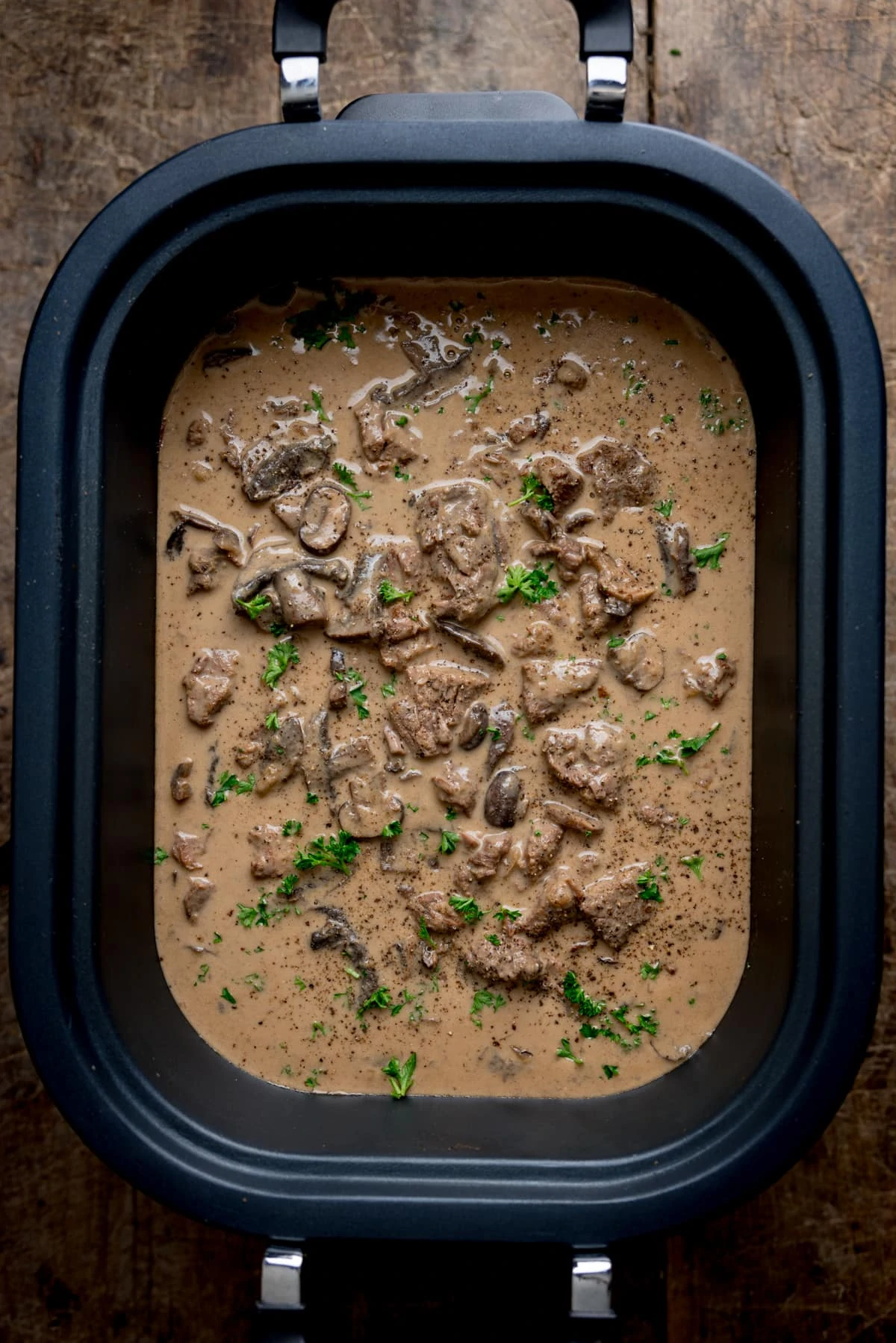 Overhead image of slow cooked beef stroganoff in a slow cooker, on a wooden table. There is parsley sprinkled on top.