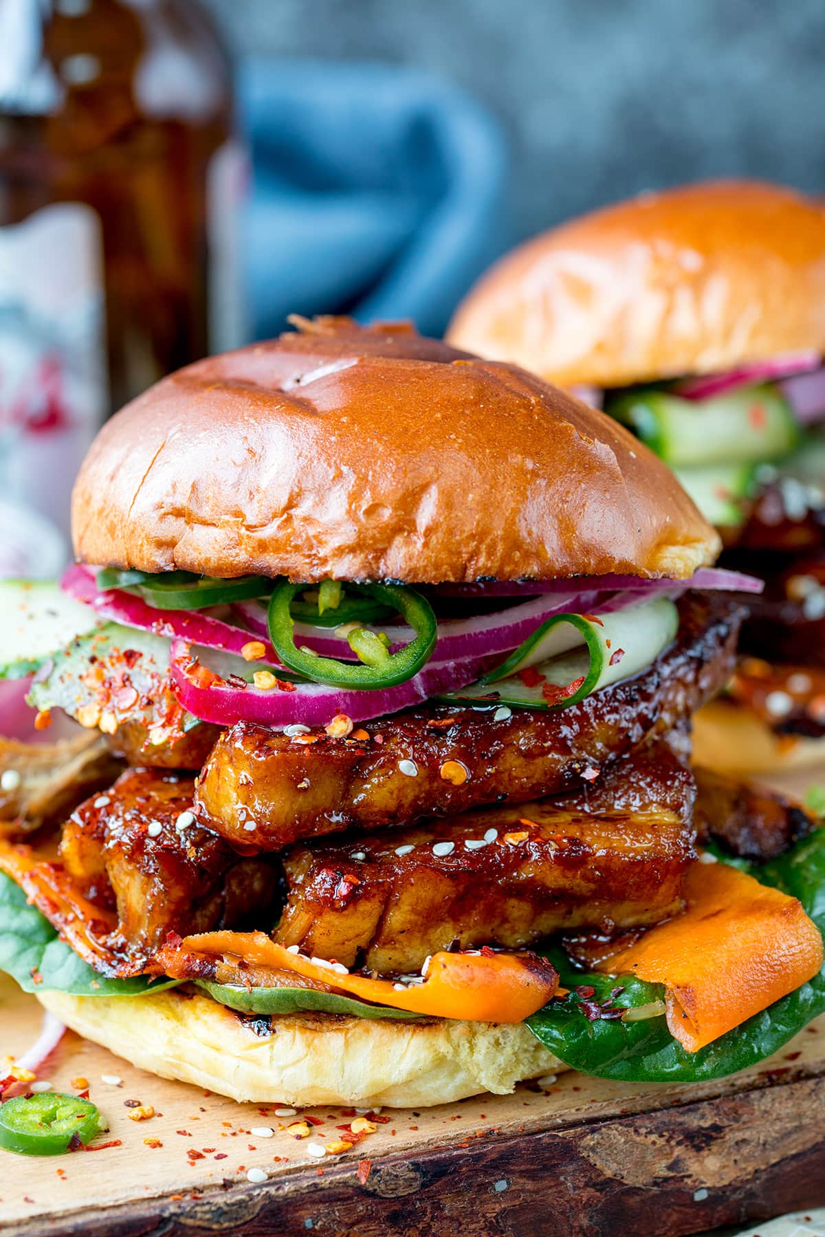 Side on image of a sticky pork belly burger with carrot, spinach, red onion and jalapeños on a brioche bun. The burger is on a wooden board and there is a further burger in the background, just in shot.