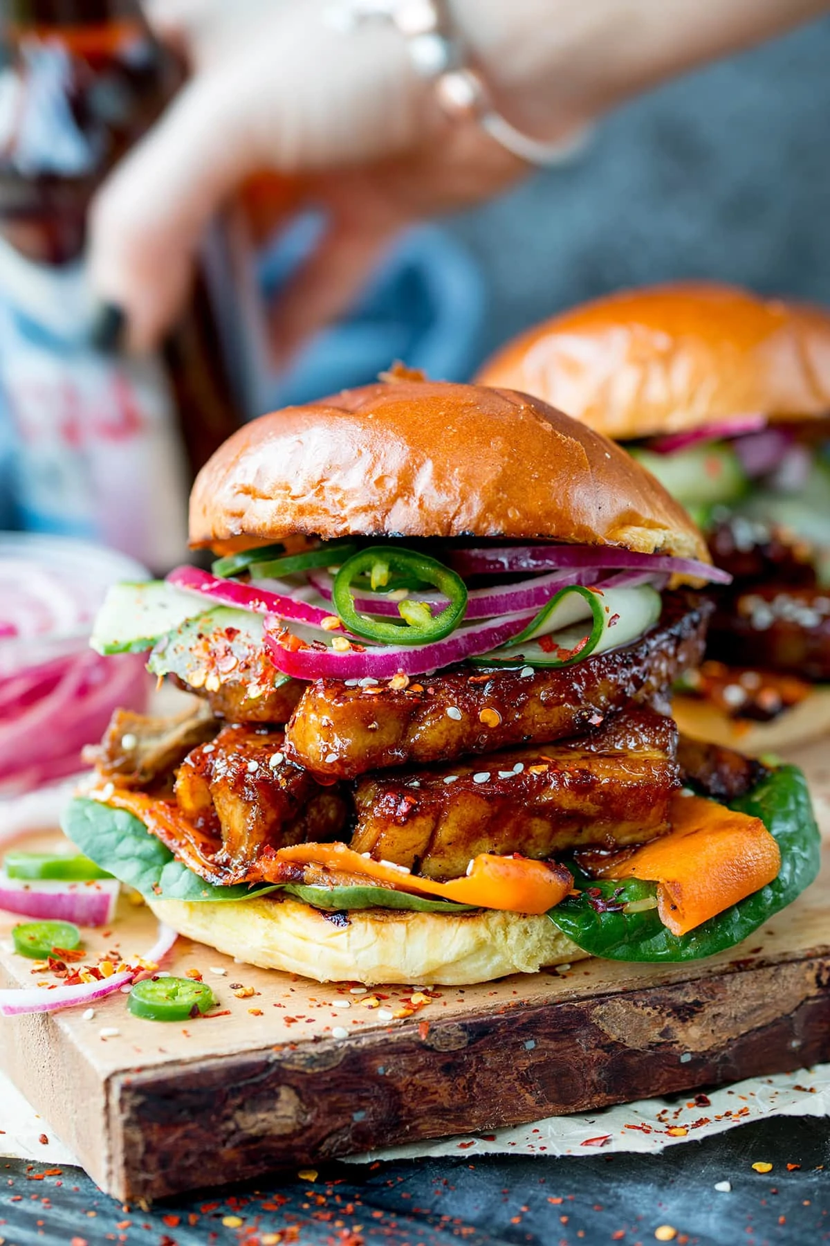 Side on image of a sticky pork belly burger with carrot, spinach, red onion and jalapeños on a brioche bun. The burger is on a wooden board and there is a further burger in the background, just in shot.
There is also a hand picking up a bottle of beer in the rear of the shot.