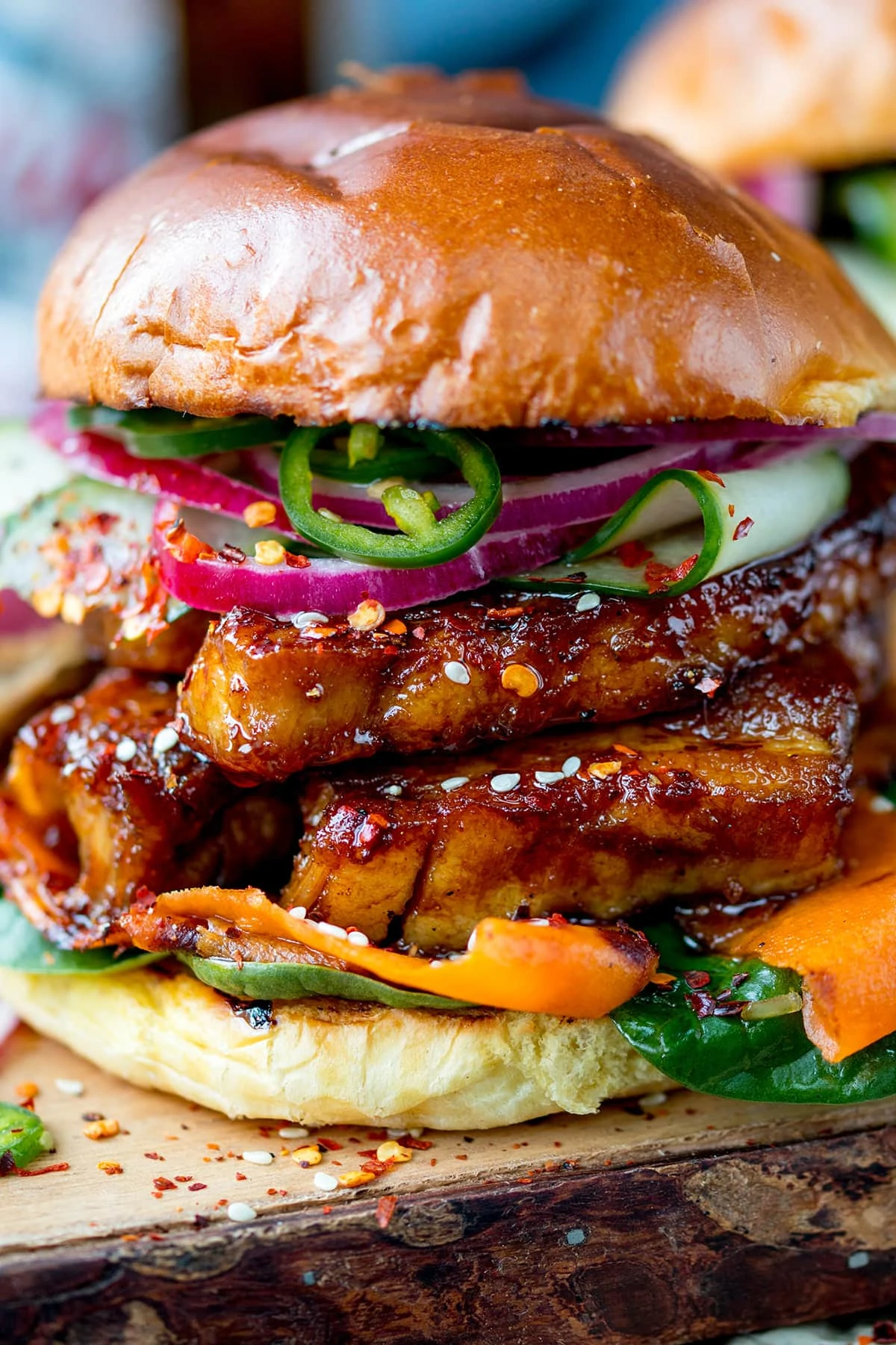 Side on, very close up image of a sticky pork belly burger with carrot, spinach, red onion and jalapeños on a brioche bun. The burger is on a wooden board. 