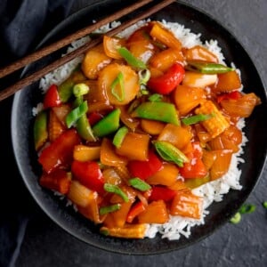 Square image of sweet and sour vegetables on a bed of boiled rice in a black bowl. There are chopsticks resting on the bowl.