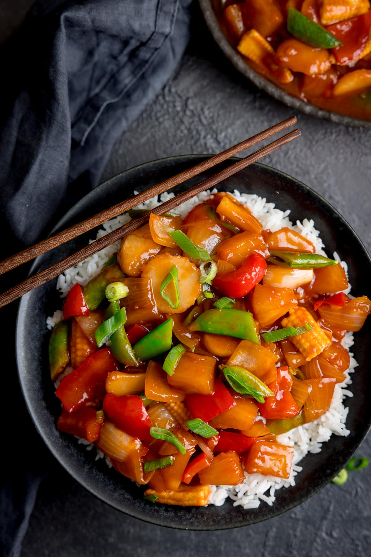 Sweet and sour vegetables on a bed of rice in a black bowl on a dark surface. There is a further bowl of sweet and sour vegetables just in shot at the top of the image.