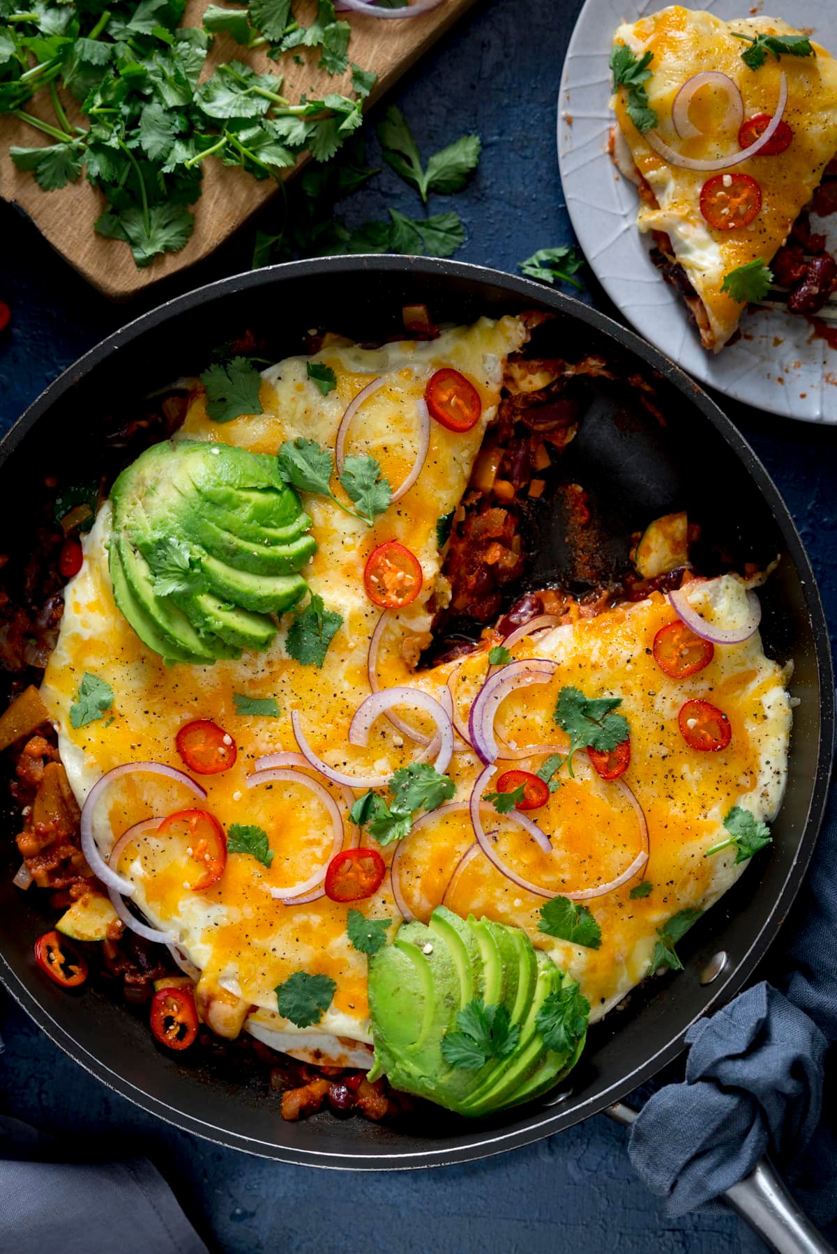 Lasagne-style chorizo, bean and tortilla pan, topped with fanned avocado, sliced red onion and red chillies. There are ingredients scattered around. A slice has been taken from the dish and is on a grey plate at the top of the image.