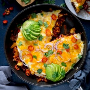 Square image of a chorizo and bean layered tortilla pan with a slice taken out. The dish is topped with sliced avocado, chopped chillies and sliced red onion.