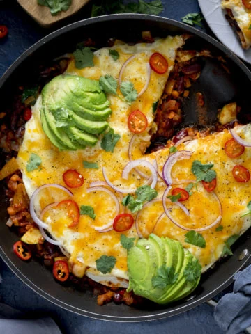 Square image of a chorizo and bean layered tortilla pan with a slice taken out. The dish is topped with sliced avocado, chopped chillies and sliced red onion.