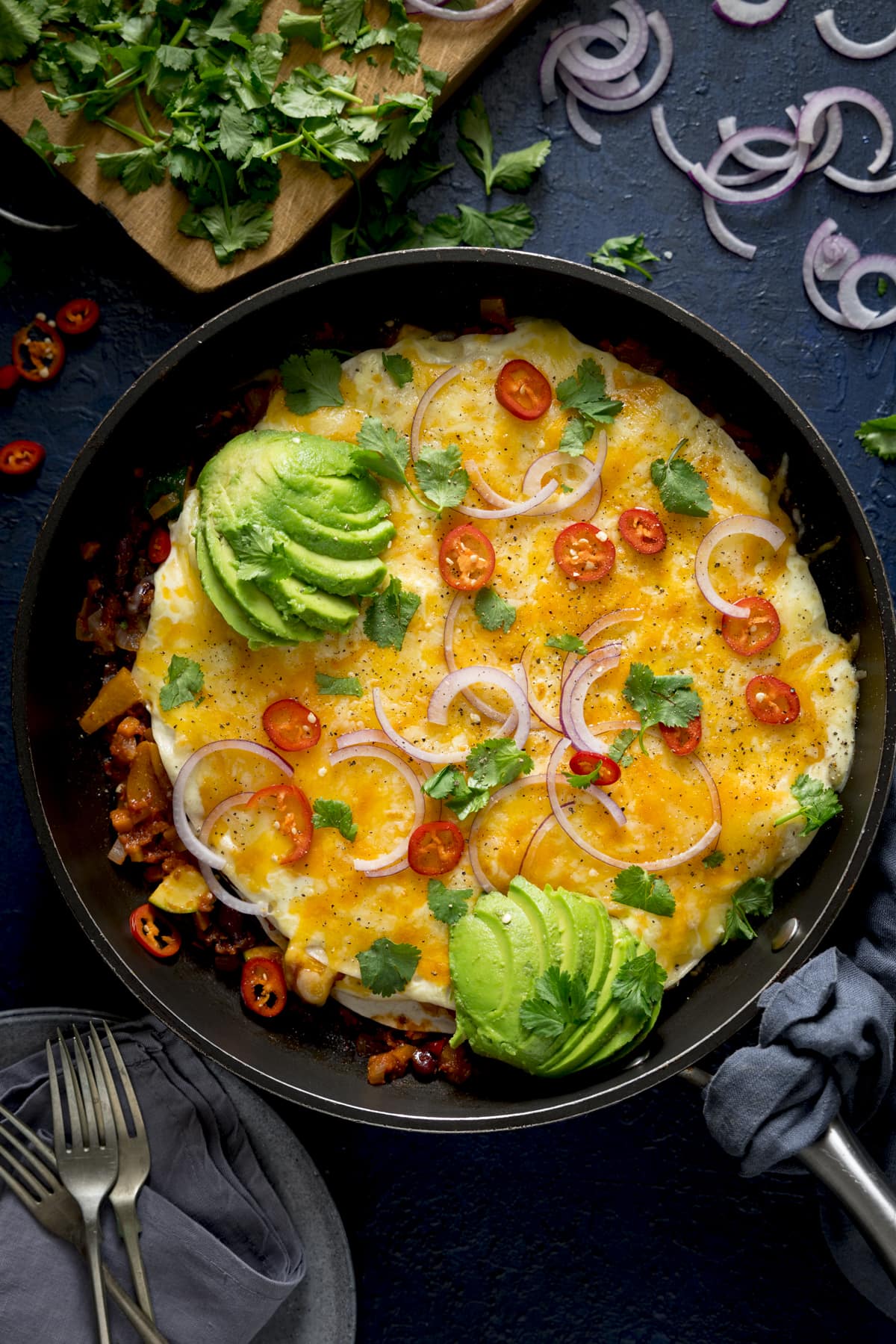Overhead image of a chorizo, bean and tortilla pan topped with melted cheese, avocado, chillies and red onion. There are ingredients scattered around the pan and the pan is on a dark background.