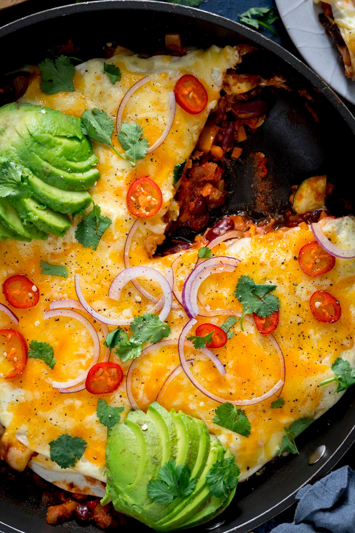 Close up overhead image of a layered chorizo, bean and tortilla pan, topped with avocado, chillies and red onion. A slice has been taken out.