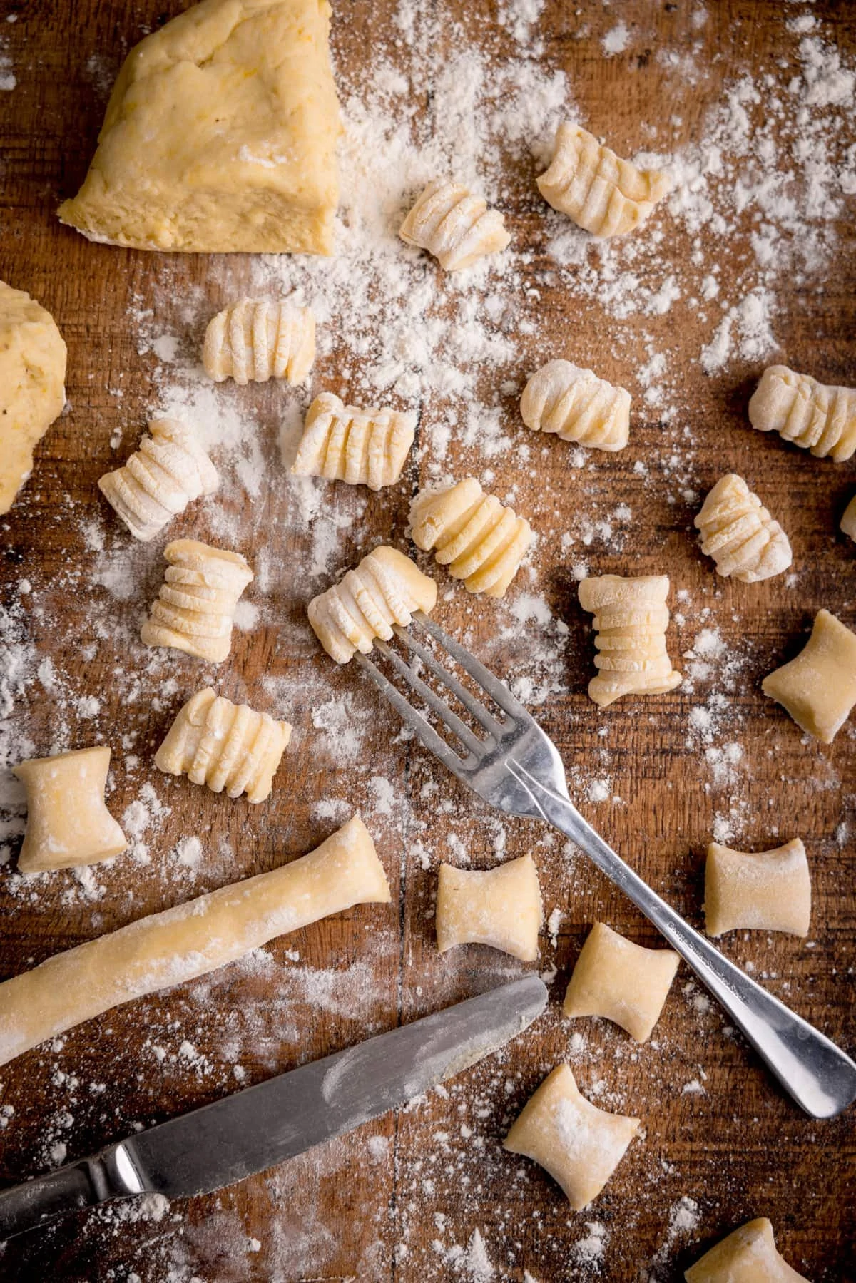 Tall image of Gnocchi being cut and shaped on a wooden board. There is a fork in shot - used for rolling into a ridged shape.
