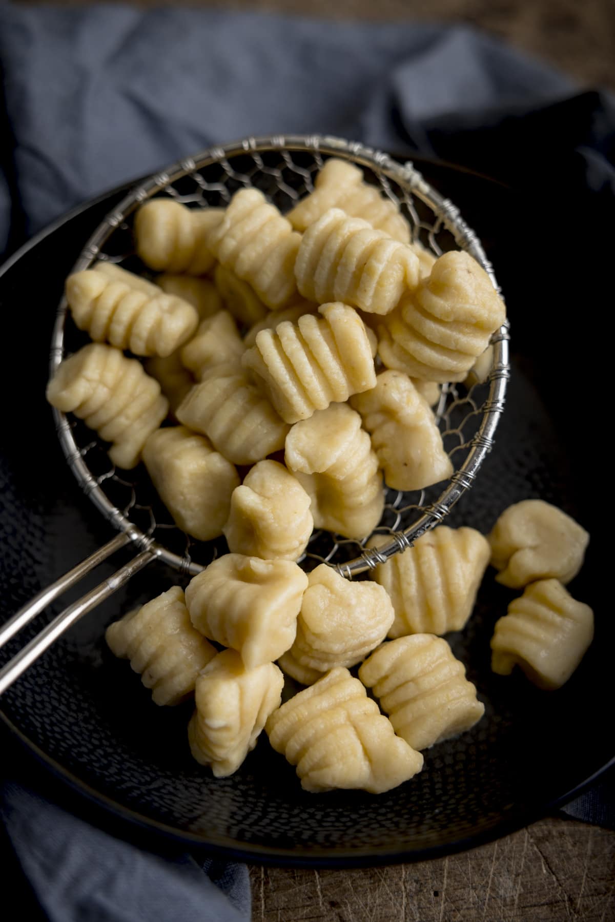 Homemade gnocchi in a slotted spoon, being added to a dark bowl. There is a blue napkin surrounding the bowl.