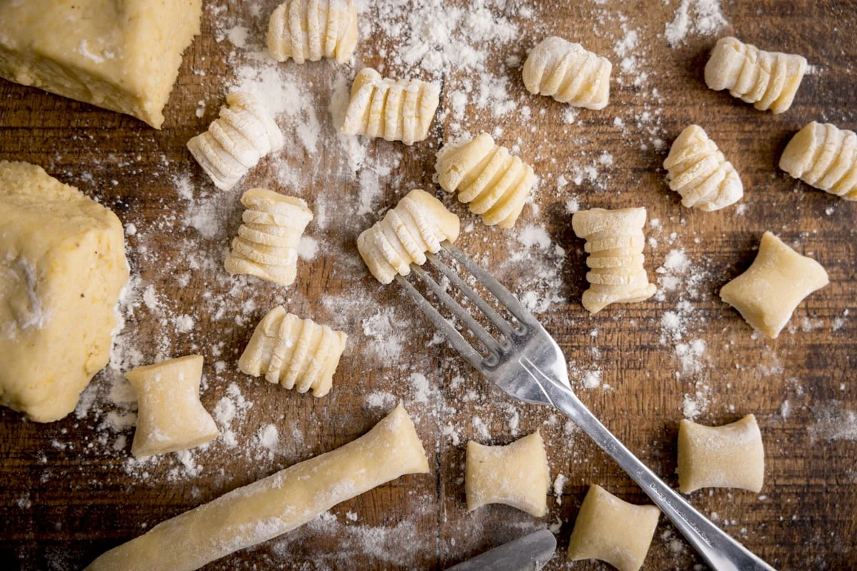 Wide image of Gnocchi being cut and shaped on a wooden board. There is a fork in shot - used for rolling.