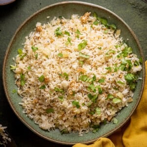 Gebratener Reis mit Zwiebeln auf einem grünen Teller mit einem senffarbenen Tuch in der unteren rechten Ecke.